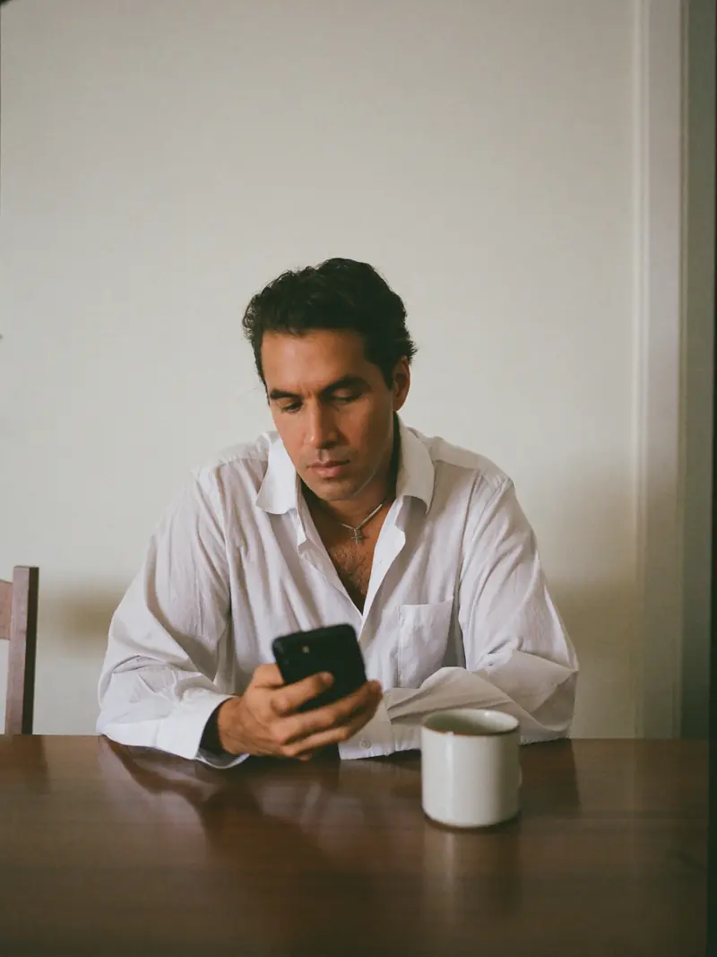 A man at his dining table in morning light, phone in hand, thinking.